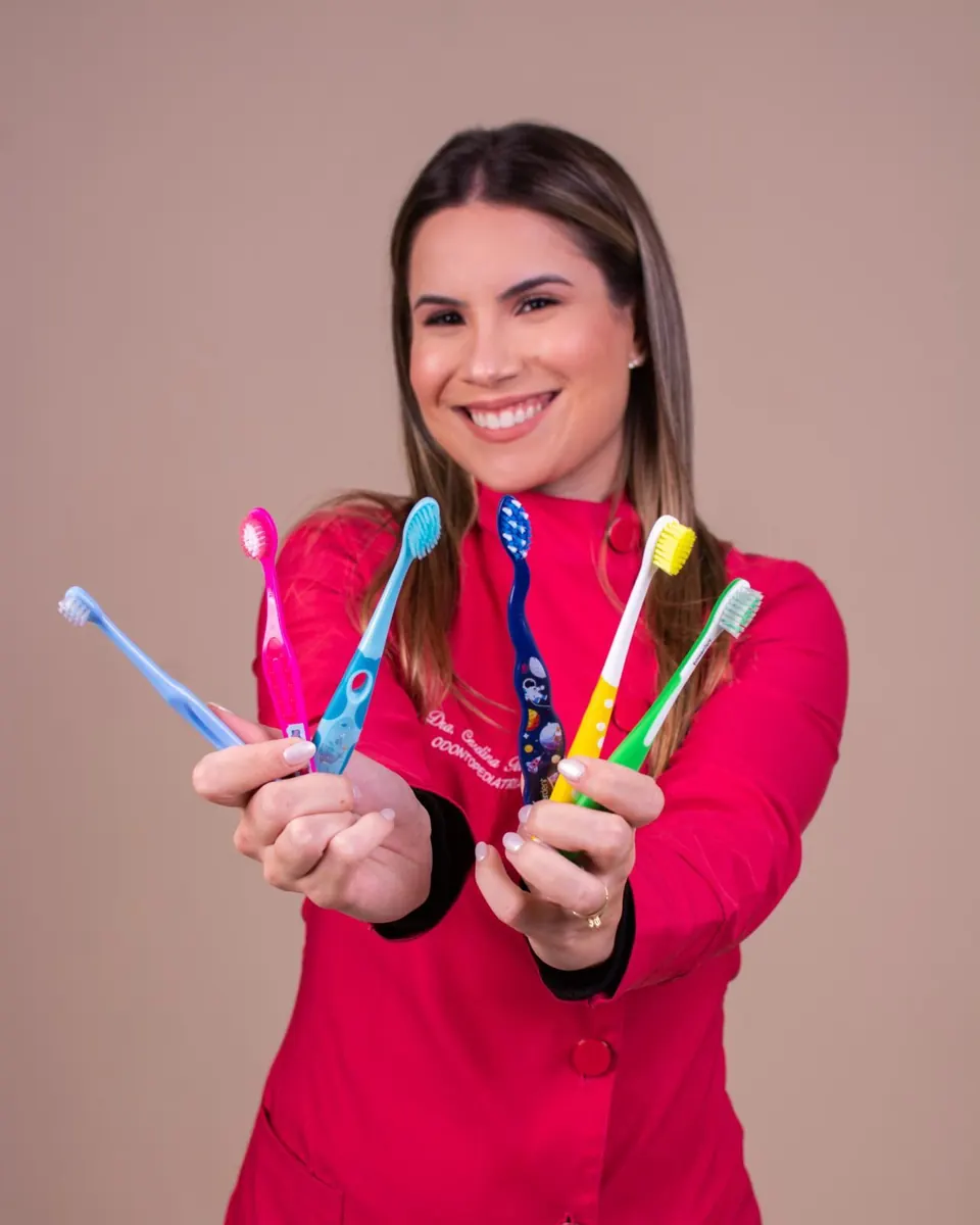 Dra. Carolina Toledo de jaleco rosa, sorrindo com escovas de dente infantis coloridas nas mãos