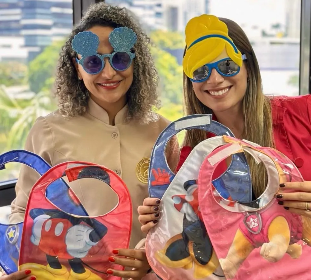 Dra. Carolina e a assistente sorrindo com óculos divertidos e babadores temáticos nas mãos